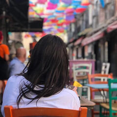A dark-haired woman wearing a white t-shirt sits outside a restaurant on a cobble street. She is facing away from the camera and the street is decorated with colourful furniture and a canopy of rainbow umbrellas.